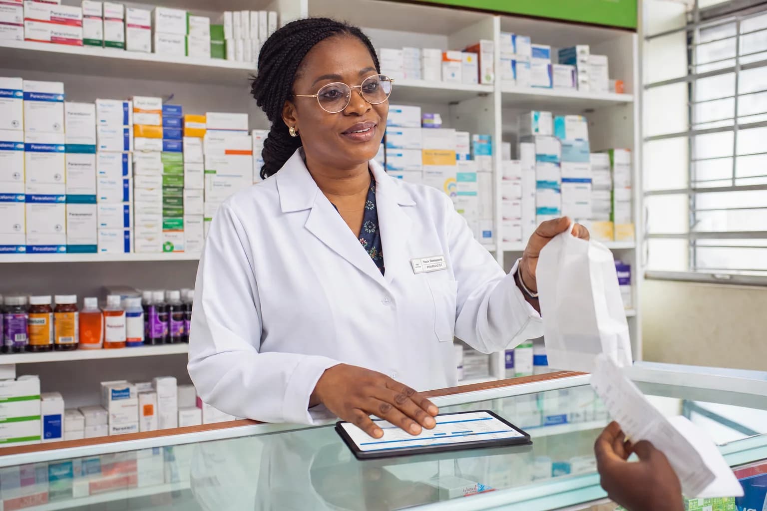 A clean small community pharmacy: a pharmacist in a white coat stands behind a glass counter, one hand on a tablet showing stock and expiry data, the other gently offering a small white bag to a customer.